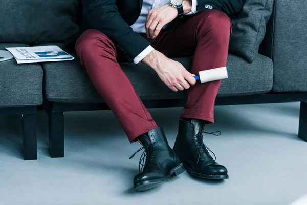 Partial view of businessman cleaning pants with sticky brush while sitting on sofa — Stock Photo