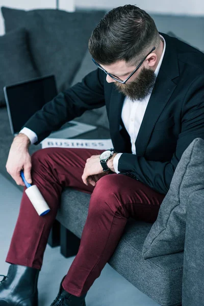 Geschäftsmann putzt Hose mit klebriger Bürste, während er auf Sofa sitzt — Stockfoto