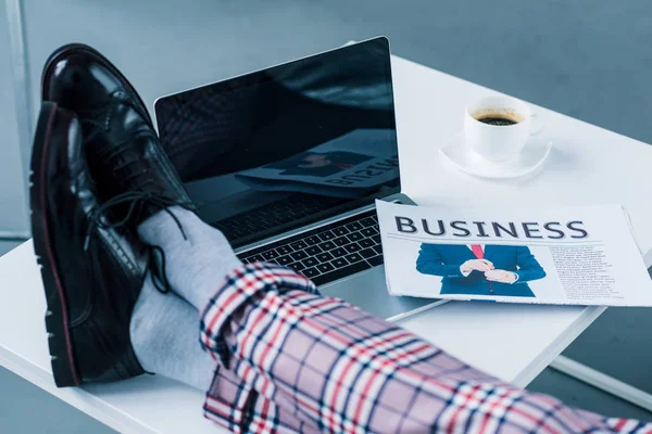 Vue partielle de l'homme d'affaires avec les jambes sur la table avec ordinateur portable et journal d'affaires — Photo de stock