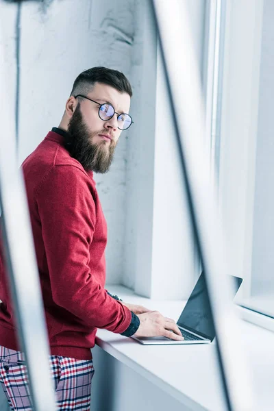 Vista lateral de hombre de negocios elegante mirando a la cámara mientras escribe en el ordenador portátil - foto de stock