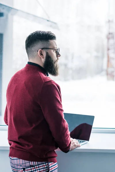 Pensive businessman in eyeglasses looking out window while working on laptop — Stock Photo
