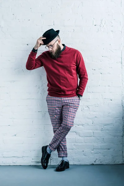 Elegante uomo barbuto in occhiali da vista con cappello in piedi contro muro di mattoni bianchi — Foto stock