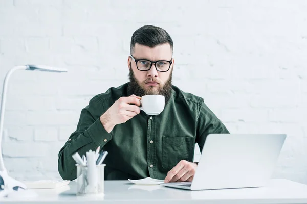 Retrato de empresario serio con taza de café en el lugar de trabajo con el ordenador portátil - foto de stock