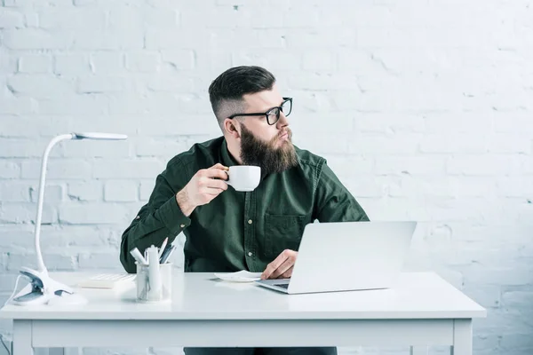 Portrait of pensive businessman with cup of coffee sitting at workplace with laptop — Stock Photo