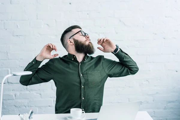 Portrait of tired businessman stretching at workplace with laptop and cup of coffee — Stock Photo