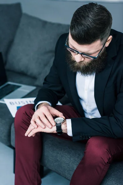 Foco seletivo do empresário verificar o tempo enquanto se senta no sofá com laptop e jornal de negócios — Fotografia de Stock
