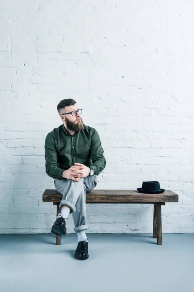 Homem de negócios pensativo sentado no banco de madeira com chapéu contra parede de tijolo branco — Fotografia de Stock