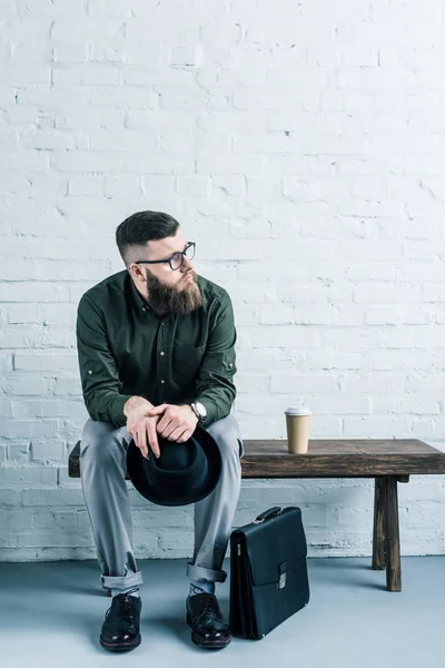 Замислений бізнесмен, з капелюхом у руках, сидячи на лавці дерев'яні проти білих цегляна стіна — стокове фото
