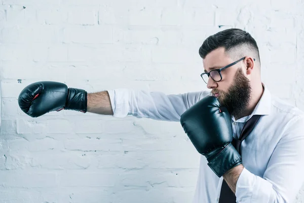 Side view of bearded businessman in eyeglasses and boxing gloves against white brick wall — Stock Photo
