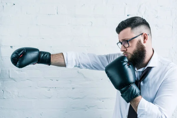 Vista lateral de hombre de negocios barbudo en gafas y guantes de boxeo contra pared de ladrillo blanco - foto de stock