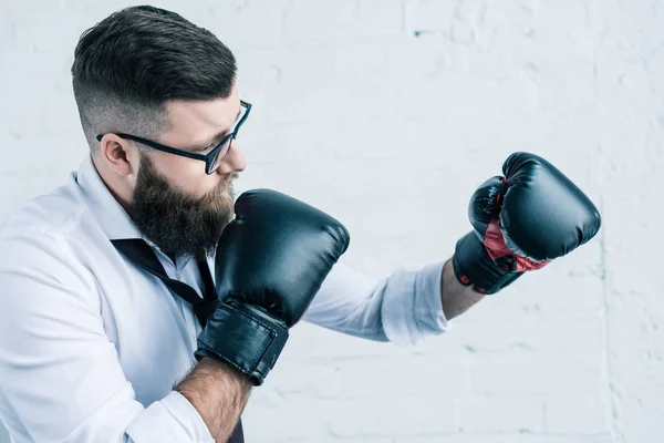 Vista laterale di uomo d'affari barbuto in occhiali e guanti da boxe contro muro di mattoni bianchi — Foto stock