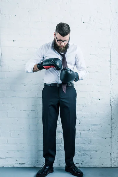 Barbudo hombre de negocios en guantes de boxeo atar pajarita contra la pared de ladrillo blanco - foto de stock
