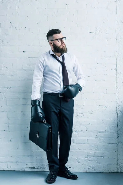 Uomo d'affari barbuto in guanti da boxe tenendo valigia contro muro di mattoni bianchi — Foto stock