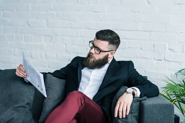 Portrait of stylish businessman in eyeglasses resting on sofa and reading newspaper — Stock Photo