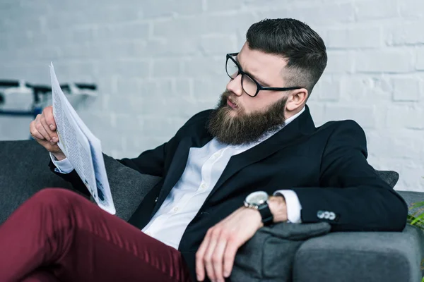 Elegante uomo d'affari barbuto appoggiato sul divano e leggendo giornali — Foto stock