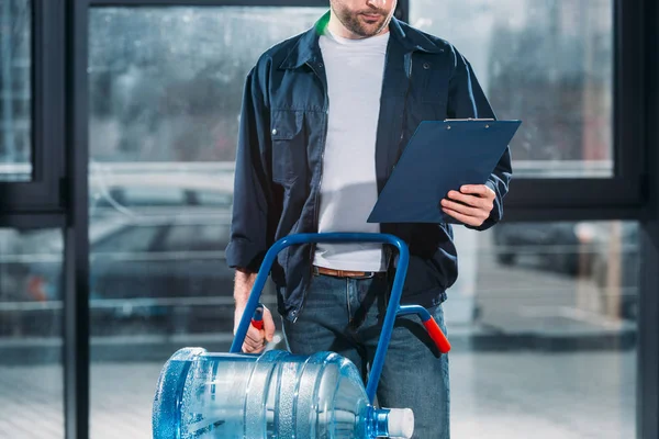 Loader looking at cargo declaration and holding delivery cart with water bottles — Stock Photo