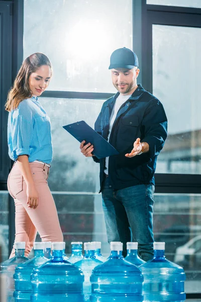 Courrier avec presse-papiers et femme regardant les bouteilles d'eau livrées — Photo de stock