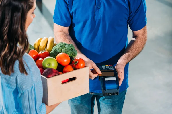 Delivery man using card and payment terminal while woman holding groceries in box — Stock Photo