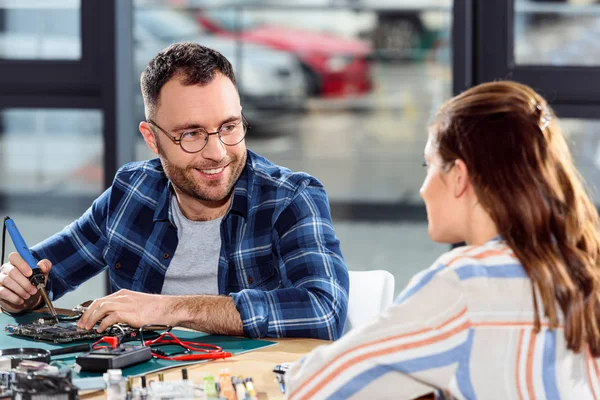 Smiling engineer soldering hardware and looking at female assistant — Stock Photo