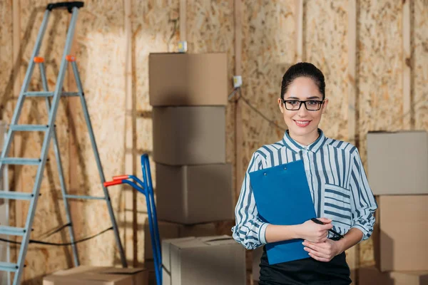 Усміхнена бізнес-леді тримає буфер обміну, що стоїть біля вантажних коробок — стокове фото