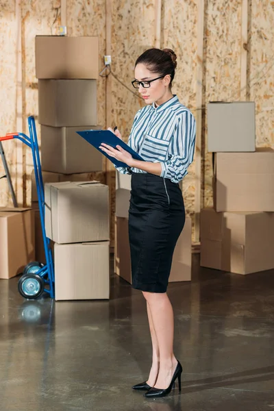 Young businesswoman writing in clipboard by stacks of delivery boxes — Stock Photo