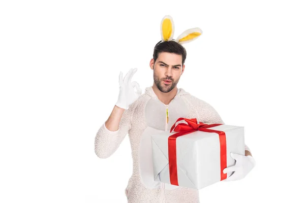 Hombre sonriente en traje de conejo sosteniendo caja de regalo y mostrando signo ok aislado en blanco, concepto de Pascua - foto de stock