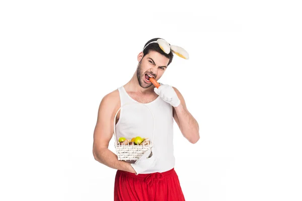 Jovem com orelhas de coelho mordendo cenoura e segurando cesta de frutas isoladas em branco, conceito de Páscoa — Fotografia de Stock