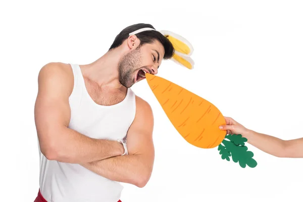 Hombre con orejas de conejo mordiendo zanahoria de la mano femenina aislado en blanco, concepto de Pascua - foto de stock