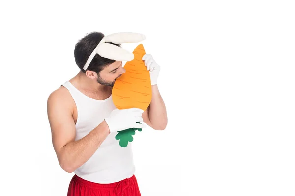 Hombre con orejas de conejo mordiendo zanahoria grande aislado en blanco, concepto de Pascua - foto de stock