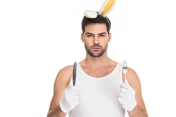 Retrato de un joven con orejas de conejo sosteniendo tenedor con cuchillo aislado en el concepto de Pascua blanca - foto de stock