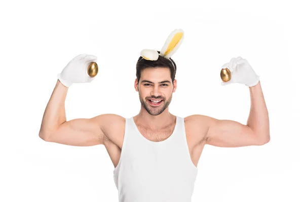 Человек с кроличьими ушами показывает мышцы и держит золотые яйца изолированы на белом, пасхальная концепция — стоковое фото