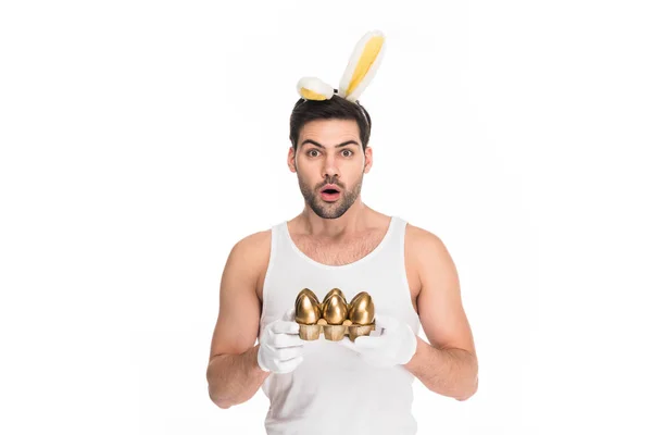 Homme choqué avec des oreilles de lapin tenant des œufs d'or isolés sur blanc, concept de Pâques — Photo de stock