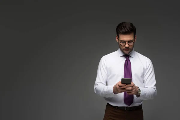 Молодой бизнесмен в очках с помощью смартфона, изолированного на сером — стоковое фото