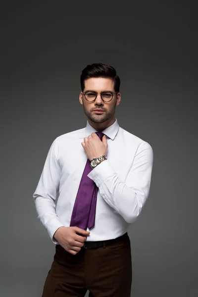 Joven empresario en gafas corrigiendo corbata aislada en gris - foto de stock