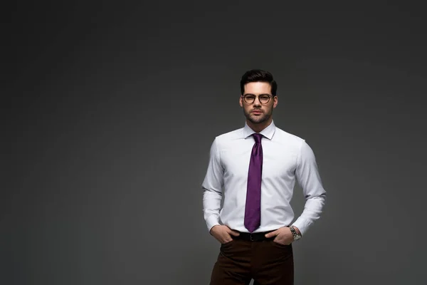 Homme d'affaires en lunettes debout avec les doigts dans des poches isolées sur gris — Photo de stock