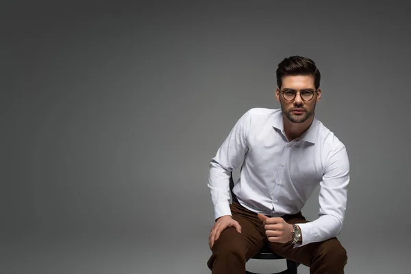 Hombre de negocios con estilo en gafas sentado en la silla y mirando a la cámara aislada en gris - foto de stock