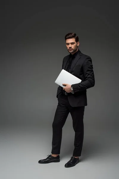 Stylish businessman standing with laptop on grey — Stock Photo