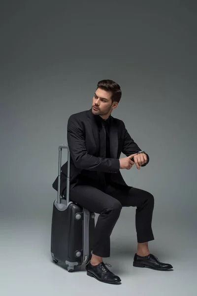 Turista de negocios sentado en la maleta y apuntando a reloj de pulsera en gris - foto de stock