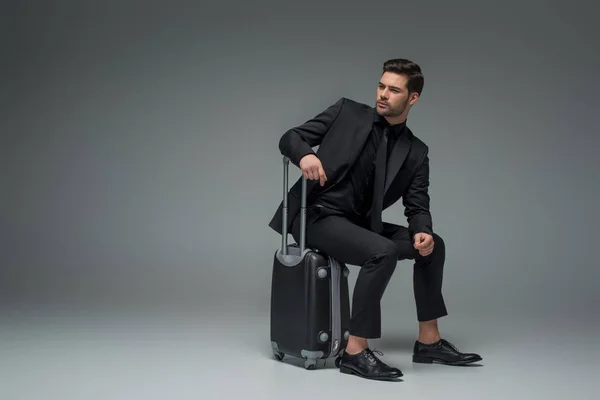 Young business tourist sitting on wheel suitcase on grey — Stock Photo