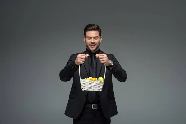 Усміхнений бізнесмен тримає кошик з фруктами ізольовано на сірому — стокове фото