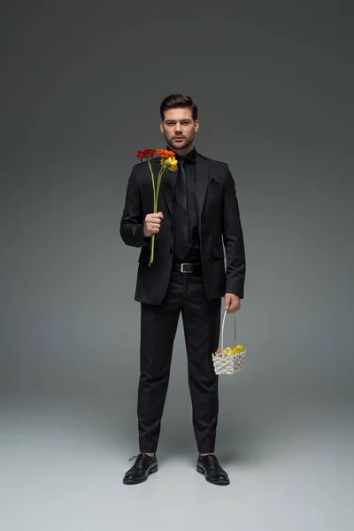 Elegante uomo d'affari che tiene fiori e cesto con frutta su grigio, concetto giornata internazionale delle donne — Foto stock