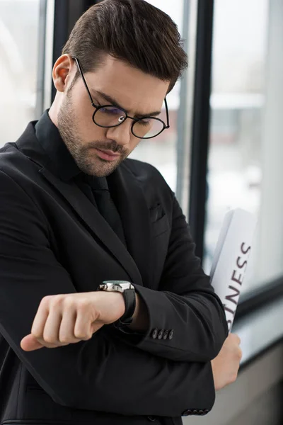 Joven hombre de negocios mirando el reloj de pulsera y sosteniendo el periódico - foto de stock