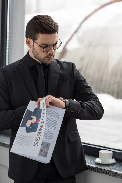 Joven hombre de negocios con gafas mirando el reloj de pulsera y sosteniendo el periódico - foto de stock