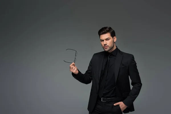 Joven hombre con estilo en traje formal sosteniendo gafas aisladas en gris - foto de stock