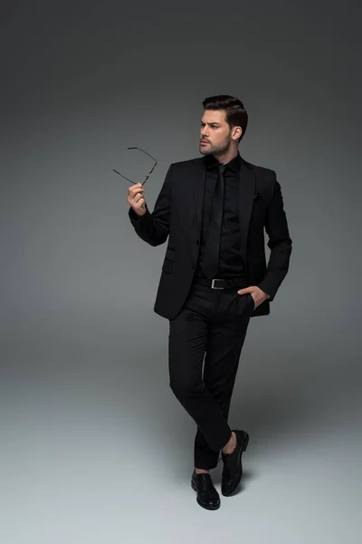 Stylish man in black suit posing with glasses in hand on grey — Stock Photo