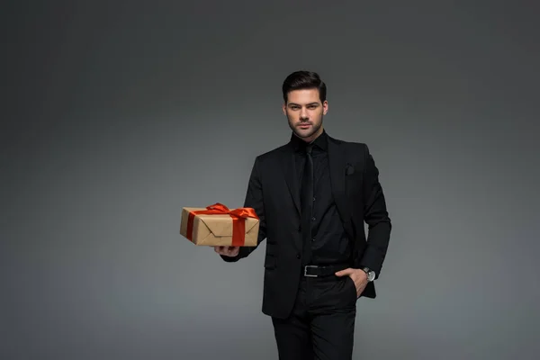 Homem elegante em terno preto segurando caixa de presente isolado em cinza, conceito de dia internacional das mulheres — Fotografia de Stock