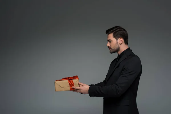 Вид збоку на стильну людину, що тримає подарункову коробку ізольовано на сірій, міжнародній концепції жіночого дня — стокове фото
