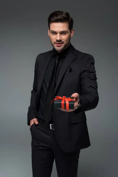 Hombre sonriente en traje negro sosteniendo caja de regalo aislado en gris, concepto de día de la mujer internacional - foto de stock