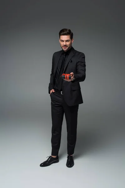 Jeune homme élégant en costume formel tenant boîte cadeau sur gris, concept de journée internationale des femmes — Photo de stock