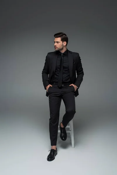 View of stylish man sitting on chair in black suit on grey — Stock Photo